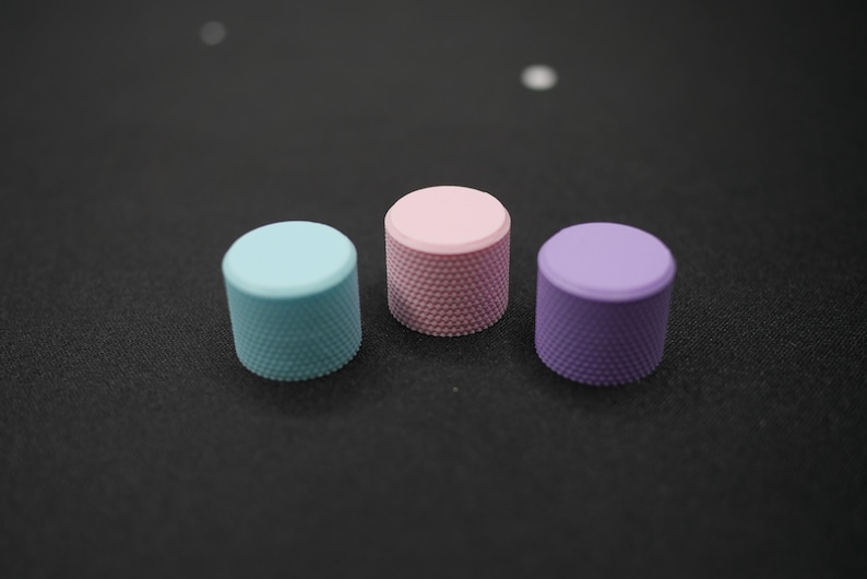 May include: Three colorful knobs in a row. The knobs are cylindrical with a textured surface. One is light blue, one is pink, and one is purple. The knobs are on a dark surface.