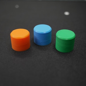 May include: Three small, cylindrical objects in orange, blue, and green. Each has a textured, dotted surface and a flat top. The objects are arranged in a row against a dark background.