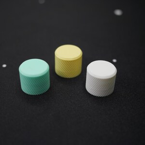 May include: Three cylindrical knobs in pastel colors: mint green, yellow, and white. Each knob has a textured, knurled surface. The knobs are arranged on a dark surface, with a few small white specks scattered around.