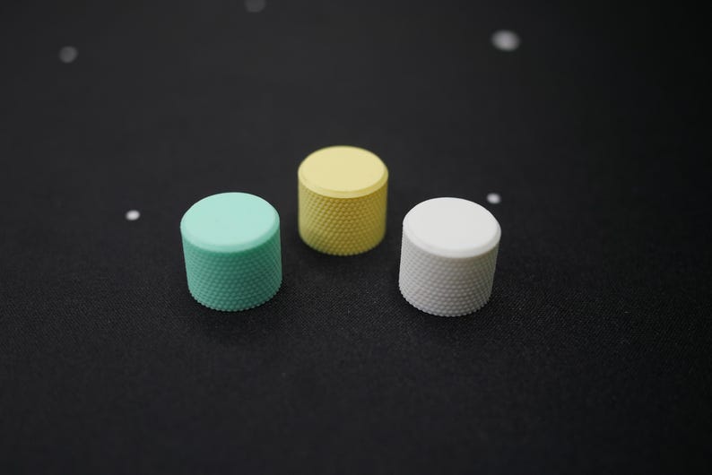 May include: Three cylindrical knobs in pastel colors: mint green, yellow, and white. Each knob has a textured, knurled surface. The knobs are arranged on a dark surface, with a few small white specks scattered around.