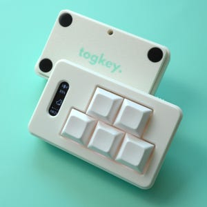 May include: Off-white Togkey keyboard with six square keys and a small display showing battery life. The top cover, also off-white, has the word "togkey." printed in a light green color. The keyboard is on a light teal background.