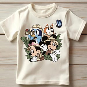 May include: A cream-colored short-sleeve t-shirt featuring a graphic print of Bluey, Bingo, Mickey Mouse, and Minnie Mouse dressed in safari gear. The characters are surrounded by green jungle leaves, a ladybug, and a blue butterfly.