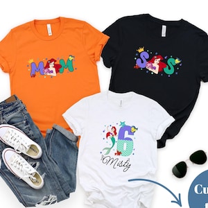 May include: Three t-shirts in orange, black, and white. The orange shirt has the word 'Mom' with a mermaid graphic. The black shirt has the letters 'S S S' with a mermaid graphic. The white shirt has the number '6' with a mermaid graphic and the name 'Misty'. Sunglasses and jeans are also present.