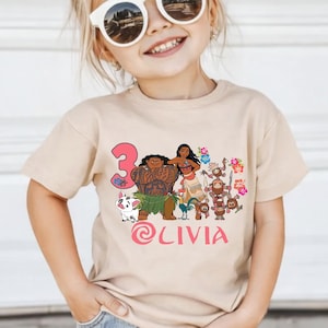 May include: A tan t-shirt featuring a colorful graphic of animated characters, the number 3, and the name "Olivia" in pink. The shirt is worn by a young girl wearing sunglasses and a flower in her hair.