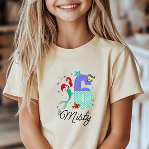 Puede incluir: Camiseta amarillo claro con un gr&aacute;fico colorido de una sirena, el n&uacute;mero 6 y el nombre "Misty". La sirena tiene el pelo rojo y una cola verde. El n&uacute;mero 6 es morado con un patr&oacute;n de escamas turquesas.