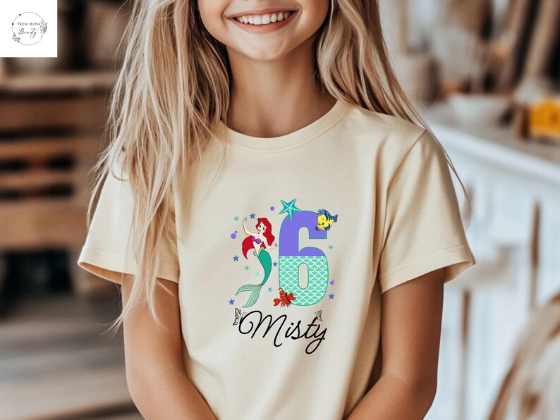 Puede incluir: Camiseta amarillo claro con un gr&aacute;fico colorido de una sirena, el n&uacute;mero 6 y el nombre "Misty". La sirena tiene el pelo rojo y una cola verde. El n&uacute;mero 6 es morado con un patr&oacute;n de escamas turquesas.