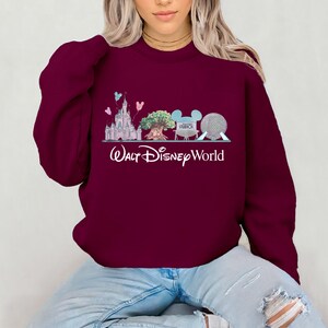 Op de afbeelding: Bordeaux sweatshirt met een afbeelding van een kasteel, boom, Mickey Mouse oren en een bol, met de woorden "Walt Disney World" in het wit. Het ontwerp bevat pastelblauwe, roze en groene accenten. Het sweatshirt is effen bordeaux.