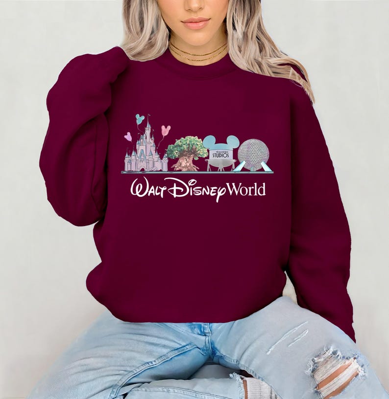 Op de afbeelding: Bordeaux sweatshirt met een afbeelding van een kasteel, boom, Mickey Mouse oren en een bol, met de woorden "Walt Disney World" in het wit. Het ontwerp bevat pastelblauwe, roze en groene accenten. Het sweatshirt is effen bordeaux.