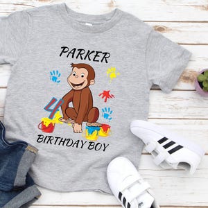 May include: A gray t-shirt with a cartoon monkey design, the name "Parker", and the text "Birthday Boy". The design includes colorful paint splatters and the number 4. White sneakers and denim shorts are also visible.