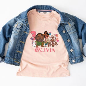 以下が含まれることがあります： 人気アニメのキャラクター、数字の3、ピンク色の「Olivia」の名前がプリントされた、明るいピーチ色のTシャツ。青いデニムジャケットの下に着ています。人気の高いアニメ映画にインスパイアされたデザインです。