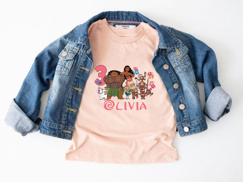 以下が含まれることがあります： 人気アニメのキャラクター、数字の3、ピンク色の「Olivia」の名前がプリントされた、明るいピーチ色のTシャツ。青いデニムジャケットの下に着ています。人気の高いアニメ映画にインスパイアされたデザインです。