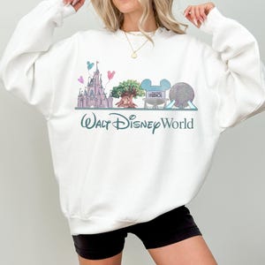 Op de afbeelding: Witte sweatshirt met een kleurrijke afbeelding van Walt Disney World-bezienswaardigheden, waaronder een kasteel, een boom en Spaceship Earth. De woorden "Walt Disney World" staan eronder in turquoise gedrukt. De sweatshirt is een comfortabele, casual stijl.