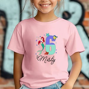 Puede incluir: Camiseta rosa con un dise&ntilde;o de sirena, el n&uacute;mero 6 y el nombre "Misty". El n&uacute;mero 6 es morado y turquesa, con una estrella y un pez. La sirena tiene el pelo rojo y una cola verde. La camiseta es para un ni&ntilde;o.