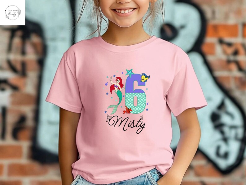 Puede incluir: Camiseta rosa con un dise&ntilde;o de sirena, el n&uacute;mero 6 y el nombre "Misty". El n&uacute;mero 6 es morado y turquesa, con una estrella y un pez. La sirena tiene el pelo rojo y una cola verde. La camiseta es para un ni&ntilde;o.