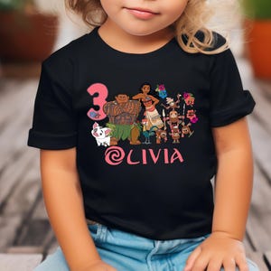 以下が含まれることがあります： アニメ映画「モアナと伝説の海」のキャラクター、モアナ、マウイ、プアなどが描かれたカラフルなグラフィックが特徴の黒いTシャツ。数字の3と名前の@LIVIAはピンク色です。子供が着ています。