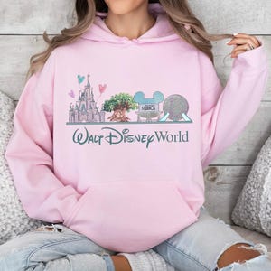 Op de afbeelding: Lichtroze hoodie met een afbeelding van het Walt Disney World-logo en parkiconen. Het ontwerp bevat een kasteel, een boom, Mickey Mouse-oren en een bol. De tekst "Walt Disney World" staat eronder gedrukt.