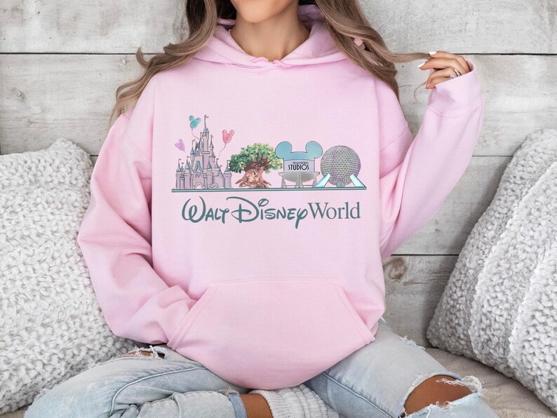Op de afbeelding: Lichtroze hoodie met een afbeelding van het Walt Disney World-logo en parkiconen. Het ontwerp bevat een kasteel, een boom, Mickey Mouse-oren en een bol. De tekst "Walt Disney World" staat eronder gedrukt.