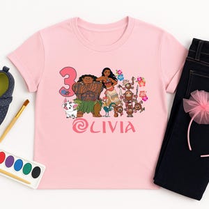 以下が含まれることがあります： ピンク色のTシャツ。映画「モアナと伝説の海」のキャラクター、数字の3、名前「LIVIA」のカラフルなグラフィックが特徴です。濃い青色のジーンズ、ピンク色のヘアバンド、濃い青色の靴も写っています。誕生日のお祝いを連想させます。