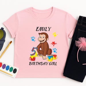 May include: A pink t-shirt with the name "Emily", a cartoon monkey, the number "5", and the words "Birthday Girl". The shirt is paired with dark blue jeans, a pink headband, and navy blue shoes.