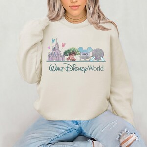 Op de afbeelding: Cr&egrave;mekleurige sweatshirt met een pastelkleurige illustratie van Walt Disney World-landmarks, waaronder een kasteel, een boom en Spaceship Earth. De woorden "Walt Disney World" zijn in een turquoise script eronder geschreven. Een casual, comfortabele stijl.