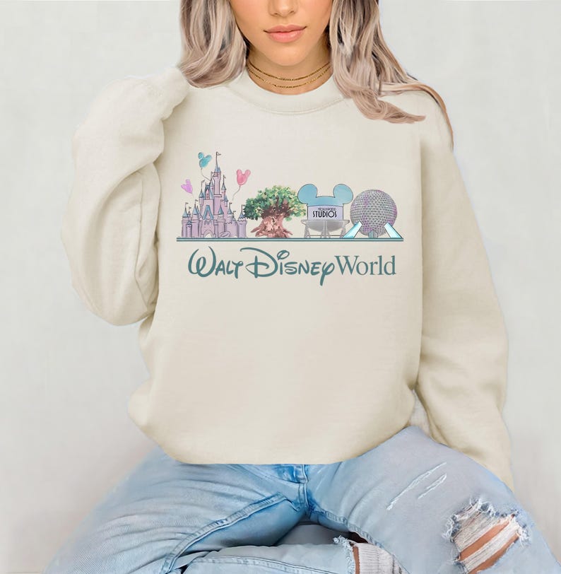 Op de afbeelding: Cr&egrave;mekleurige sweatshirt met een pastelkleurige illustratie van Walt Disney World-landmarks, waaronder een kasteel, een boom en Spaceship Earth. De woorden "Walt Disney World" zijn in een turquoise script eronder geschreven. Een casual, comfortabele stijl.