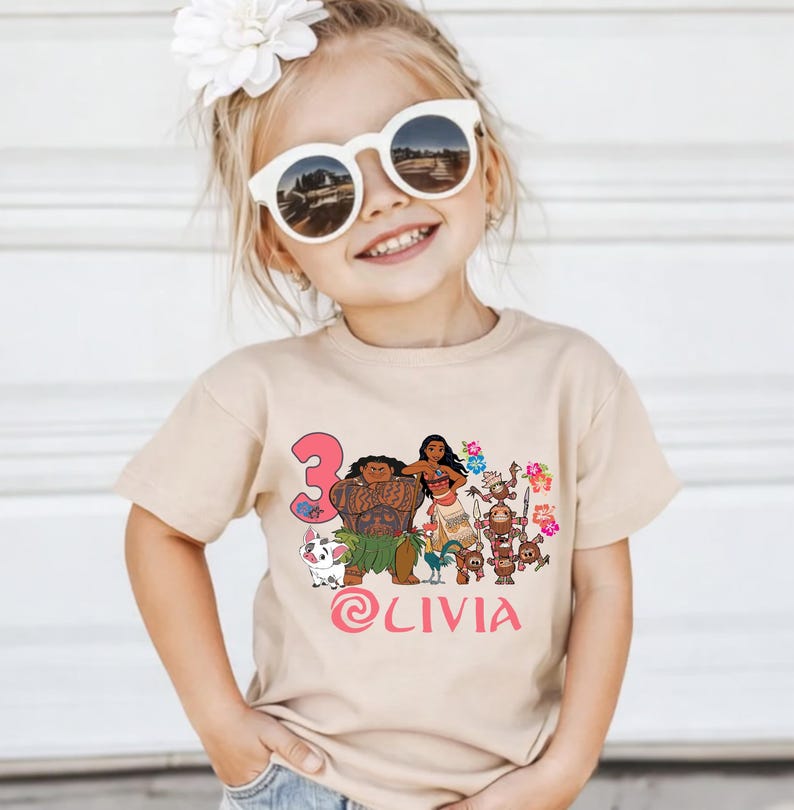 May include: A tan t-shirt featuring a colorful graphic of animated characters, the number 3, and the name "Olivia" in pink. The shirt is worn by a young girl wearing sunglasses and a flower in her hair.
