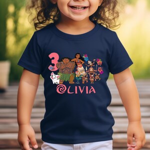 以下が含まれることがあります： ネイビーブルーの幼児用Tシャツ。人気アニメのキャラクターのカラフルなグラフィックが特徴です。「3」の数字はピンク色で、「Olivia」という名前もピンク色の筆記体で表示されています。デザインはシャツの中央に配置されています。