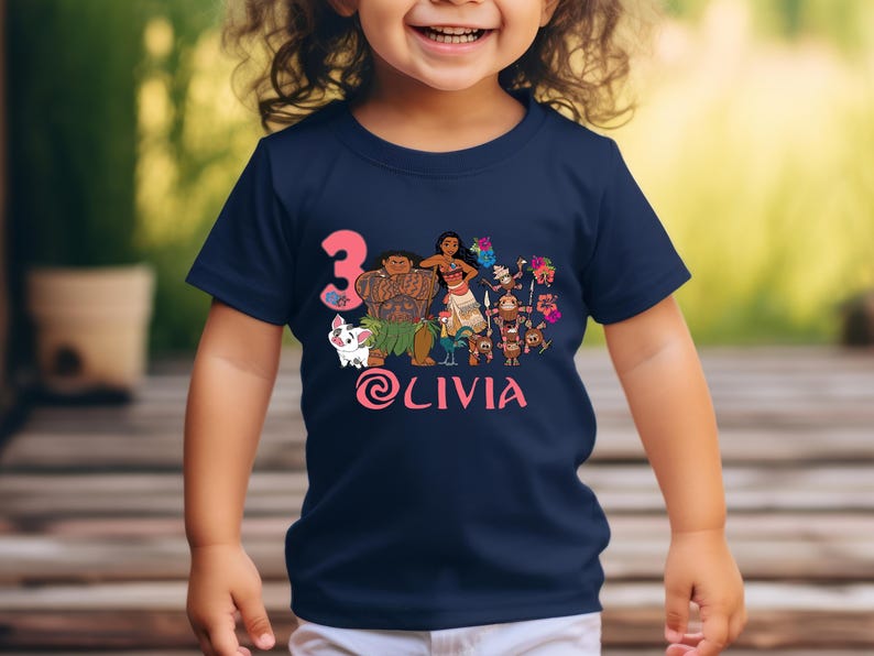 以下が含まれることがあります： ネイビーブルーの幼児用Tシャツ。人気アニメのキャラクターのカラフルなグラフィックが特徴です。「3」の数字はピンク色で、「Olivia」という名前もピンク色の筆記体で表示されています。デザインはシャツの中央に配置されています。