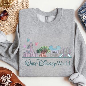 Op de afbeelding: Een grijze sweatshirt met een kleurrijke afbeelding van het Walt Disney World-logo en parkafbeeldingen. Het ontwerp omvat een kasteel, een boom en Spaceship Earth. De tekst "Walt Disney World" staat eronder.