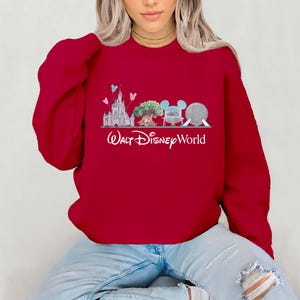Op de afbeelding: Een rode sweatshirt met een Walt Disney World-ontwerp. De afbeelding toont een kasteel, boom, Mickey Mouse-oren en een bol. De woorden "Walt Disney World" staan eronder in witte letters.
