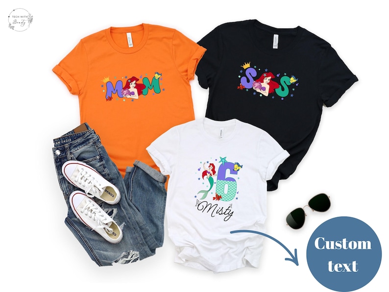 May include: Three t-shirts in orange, black, and white. The orange shirt has the word 'Mom' with a mermaid graphic. The black shirt has the letters 'S S S' with a mermaid graphic. The white shirt has the number '6' with a mermaid graphic and the name 'Misty'. Sunglasses and jeans are also present.