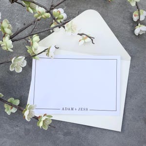 May include: A blank white card with a black border sits inside a cream-colored envelope. The text "ADAM & JESS" is printed in black at the bottom of the card. Decorative branches with white and green flowers are arranged around the card and envelope.
