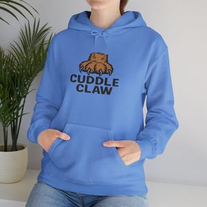 May include: A light blue hoodie with a front pocket and a drawstring hood. The hoodie features a brown bear claw graphic above the words "CUDDLE CLAW" in black. The person is wearing blue jeans.