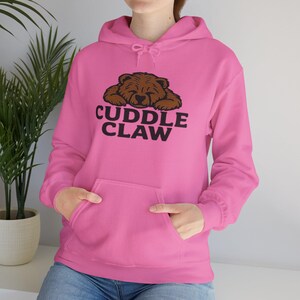 May include: A bright pink hoodie with a brown bear graphic and the words "Cuddle Claw" in black. The hoodie has a front pocket and a drawstring hood. The sleeves are long and the material appears to be a soft fabric.