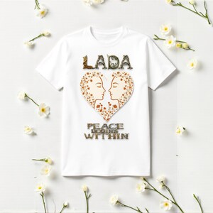 Op de afbeelding: Wit T-shirt met het woord "LADA" boven een hartvormig ontwerp met twee profielen en bloemendetails. De zin "PEACE BEGINS WITHIN" staat eronder. Het T-shirt is versierd met witte bloemen.