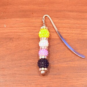 Puede incluir: Un marcapáginas de metal con una hilera vertical de cuatro cuentas esféricas texturizadas en amarillo, blanco, rosa y negro. Cada cuenta está separada por pequeños espaciadores de tono plateado con cristales transparentes incrustados.