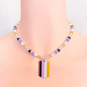 Op de afbeelding: Een kralenketting met een rechthoekige hanger in de vorm van een dog tag met verticale strepen in zwart, wit, paars en geel. De ketting is samengesteld uit afwisselende ronde kralen in paars, geel, wit en transparant met zilverkleurige tussenstukjes.