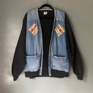 May include: A blue denim jacket with black sleeves and a black knit collar. The jacket has two large pockets on the front and two smaller pockets on the chest. The chest pockets have a geometric pattern in brown, white, and blue.