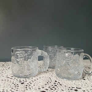 May include: Three clear glass mugs with a raised design of a cartoon character. The mugs have a handle and are perfect for a hot beverage.