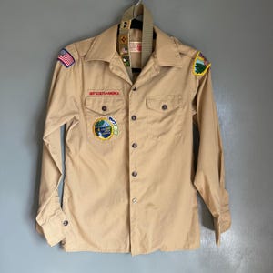 May include: Tan Boy Scouts of America shirt with long sleeves, two chest pockets, and button closures. Patches include an American flag, a landscape, and a circular emblem. A tan sash with additional patches hangs from the collar.