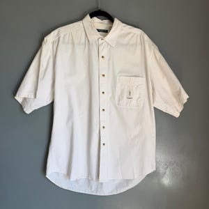 May include: Off-white short-sleeve button-down shirt with a chest pocket. The shirt has a classic collar and wooden-colored buttons. The pocket features a small logo and the word "Gant". The shirt is hanging on a black hanger.