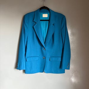 Puede incluir: Una blazer turquesa con cierre de dos botones, dos bolsillos delanteros y tres botones en cada manga. La blazer está colgada de una percha negra sobre un fondo gris. El forro interior es de un tono turquesa más claro.