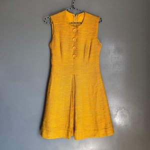 May include: A sleeveless, mustard yellow dress with a textured fabric. The dress features a round neckline, a front zipper, and three decorative buttons. The skirt has a pleated design, and the dress hangs on a black and white patterned hanger.