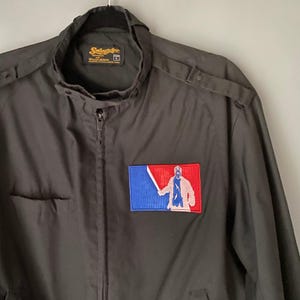 May include: A black Swingster World Wearables jacket with a large embroidered patch on the left chest. The patch features a baseball diamond with a blue, red, and white color scheme. A figure in a white shirt and black pants is holding a baseball bat. The jacket has a zipper closure and a pocket on the left side.