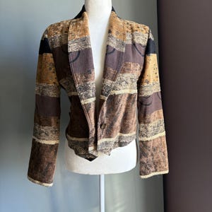 May include: A patterned blazer in shades of brown, tan, and black, displayed on a mannequin. The jacket features a notched collar, long sleeves, and a single-button closure. The fabric has a textured appearance with a geometric design.