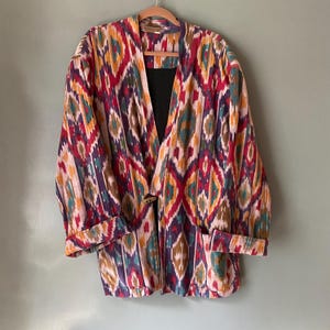 Puede incluir: Una chaqueta colorida con un diseño geométrico en tonos rojos, azules, verdes y amarillos. La chaqueta tiene un cierre de botón y un ajuste holgado.