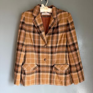 Puede incluir: Una blazer vintage a cuadros en tonos marrón, beige y negro, con un acento naranja. La blazer presenta un cuello con solapas clásico, dos bolsillos delanteros y un cierre de dos botones. La chaqueta está colgada de una percha blanca.