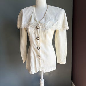 May include: Cream-colored long-sleeve blouse with a V-neck and a decorative lace collar. The blouse features three dark brown buttons down the front and is displayed on a mannequin. The fabric appears to be a lightweight material.