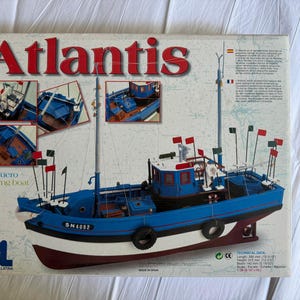 Könnte beinhalten: Ein Modellbausatz eines Fischerbootes mit dem Namen "Atlantis" in roten Buchstaben. Das Boot ist blau, weiß und rot, mit mehreren Masten und Flaggen. Die Box enthält Detailbilder des Bootes und den Text "Pesquero Fishing Boat".
