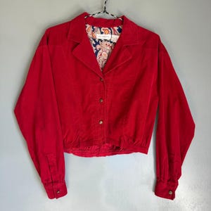 May include: A vibrant red corduroy jacket with a collared neckline and long sleeves. The jacket features a button-down front and a patterned paisley lining. The cropped style adds a vintage flair to this outerwear piece.